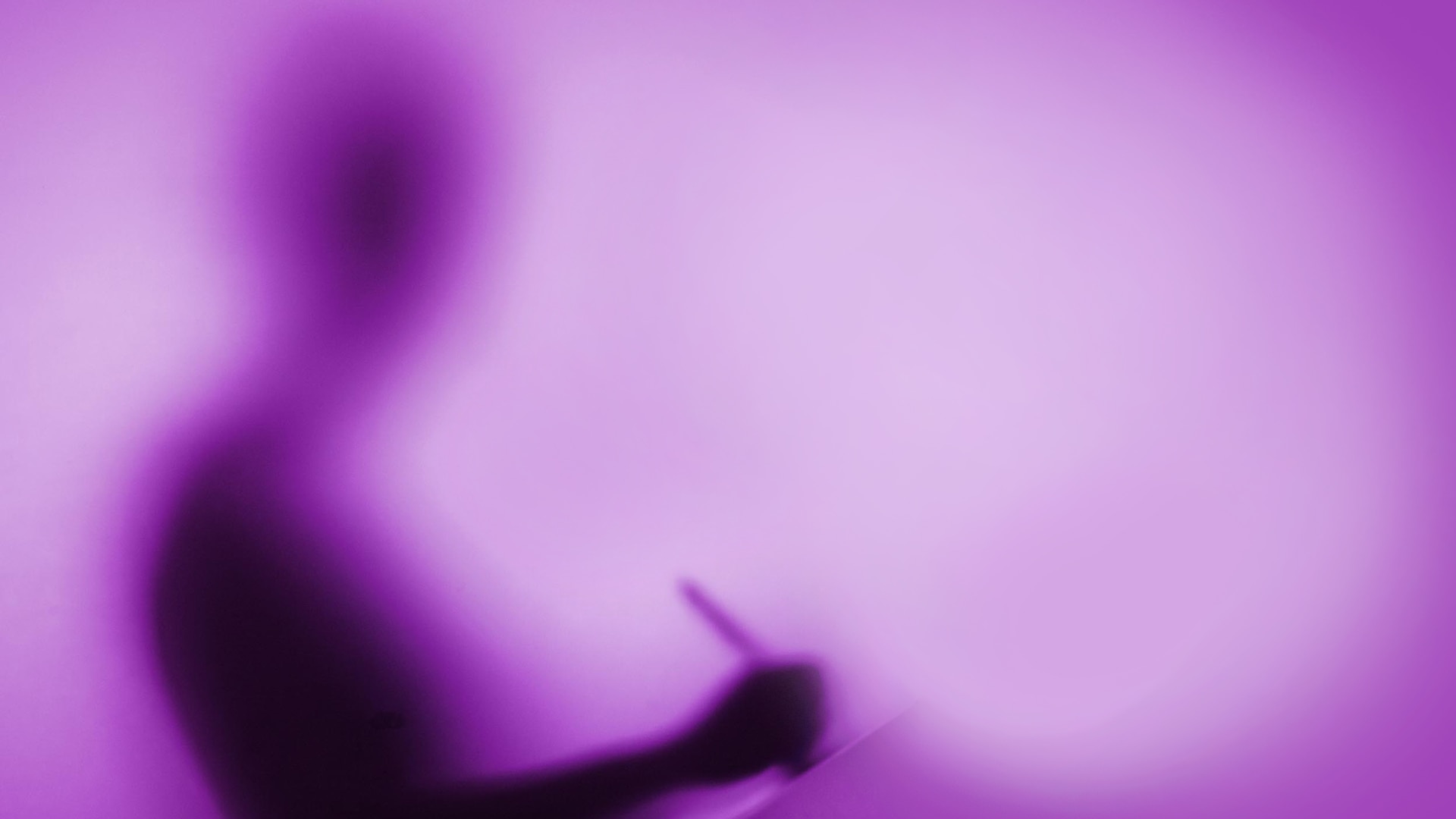   The blurry silhouette of a person writing on a pad seen against a purple background