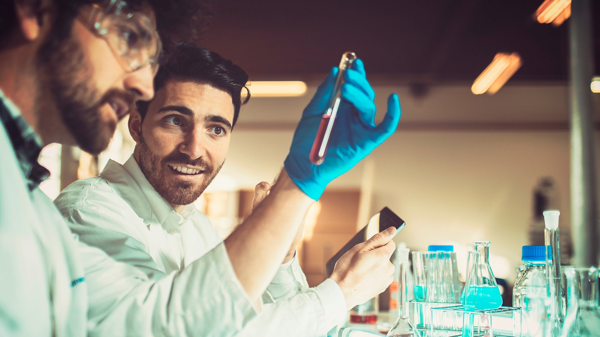    Two men in lab coats sitting at a laboratory bench look at a test tube